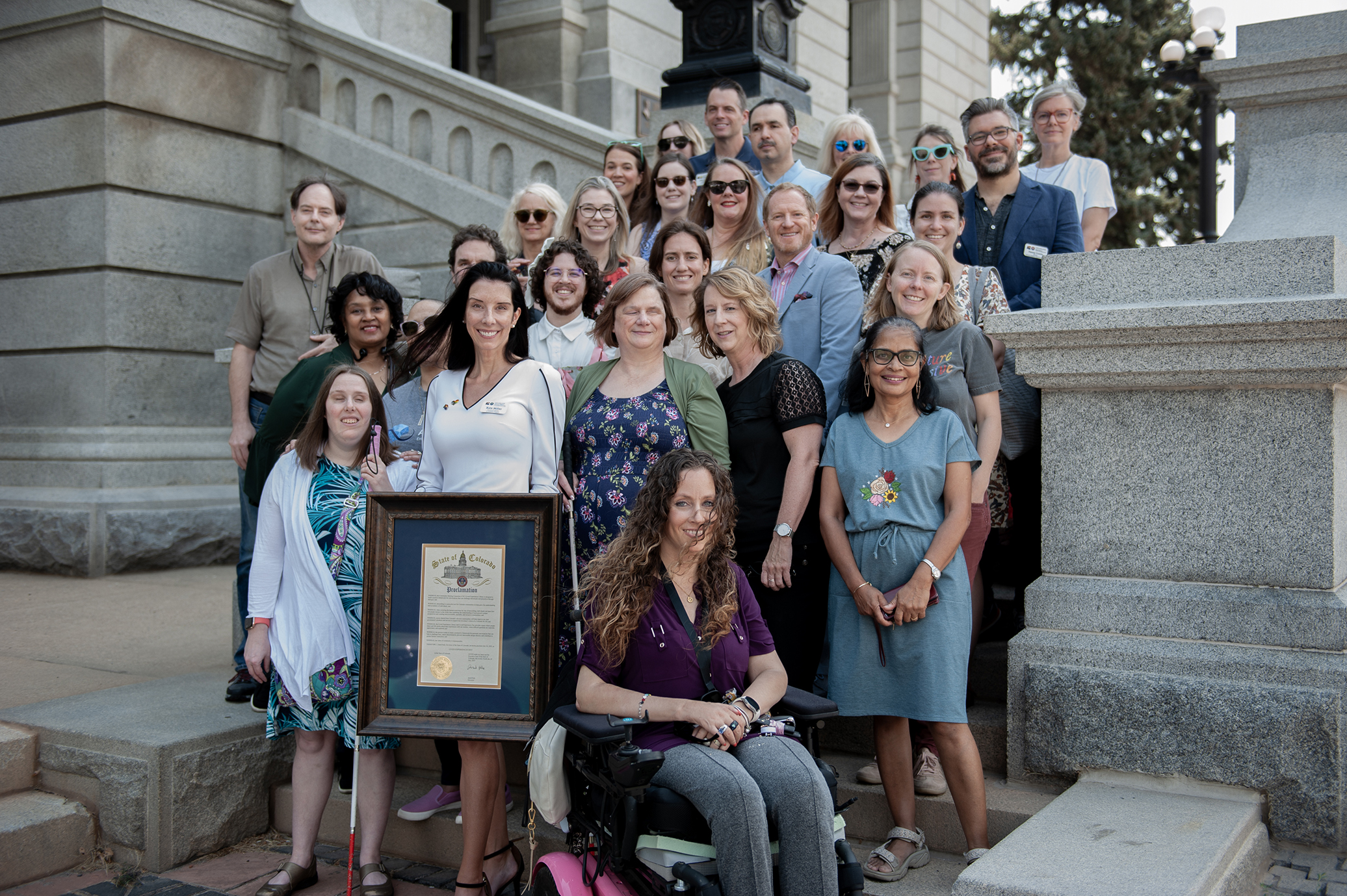 The TAP team, Community Steering Committee, and accessibility allies stand at the Colorado State Capitol steps with a framed proclamation