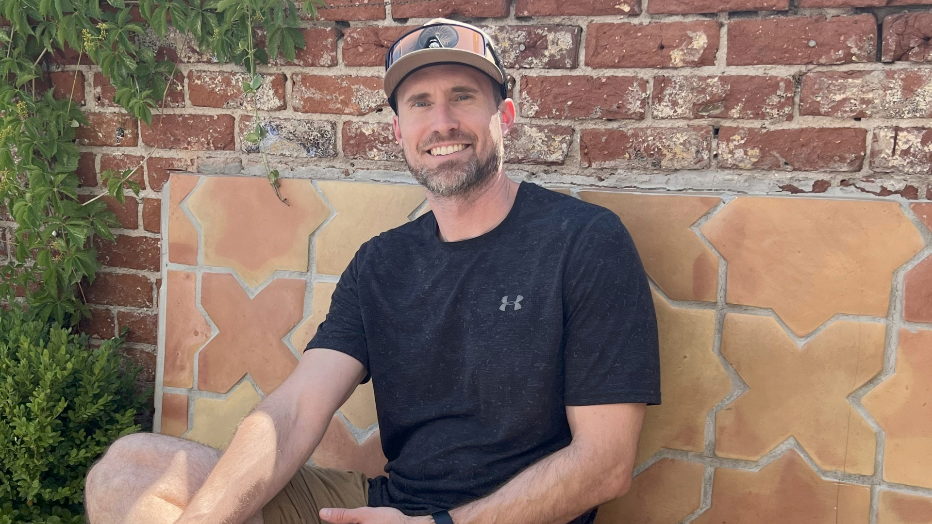 Sean Clark with a beard and a baseball cap is seated on a tiled bench outdoors, smiling at the camera. He's wearing a dark t-shirt, khaki shorts, and athletic shoes. Behind him is a brick wall.