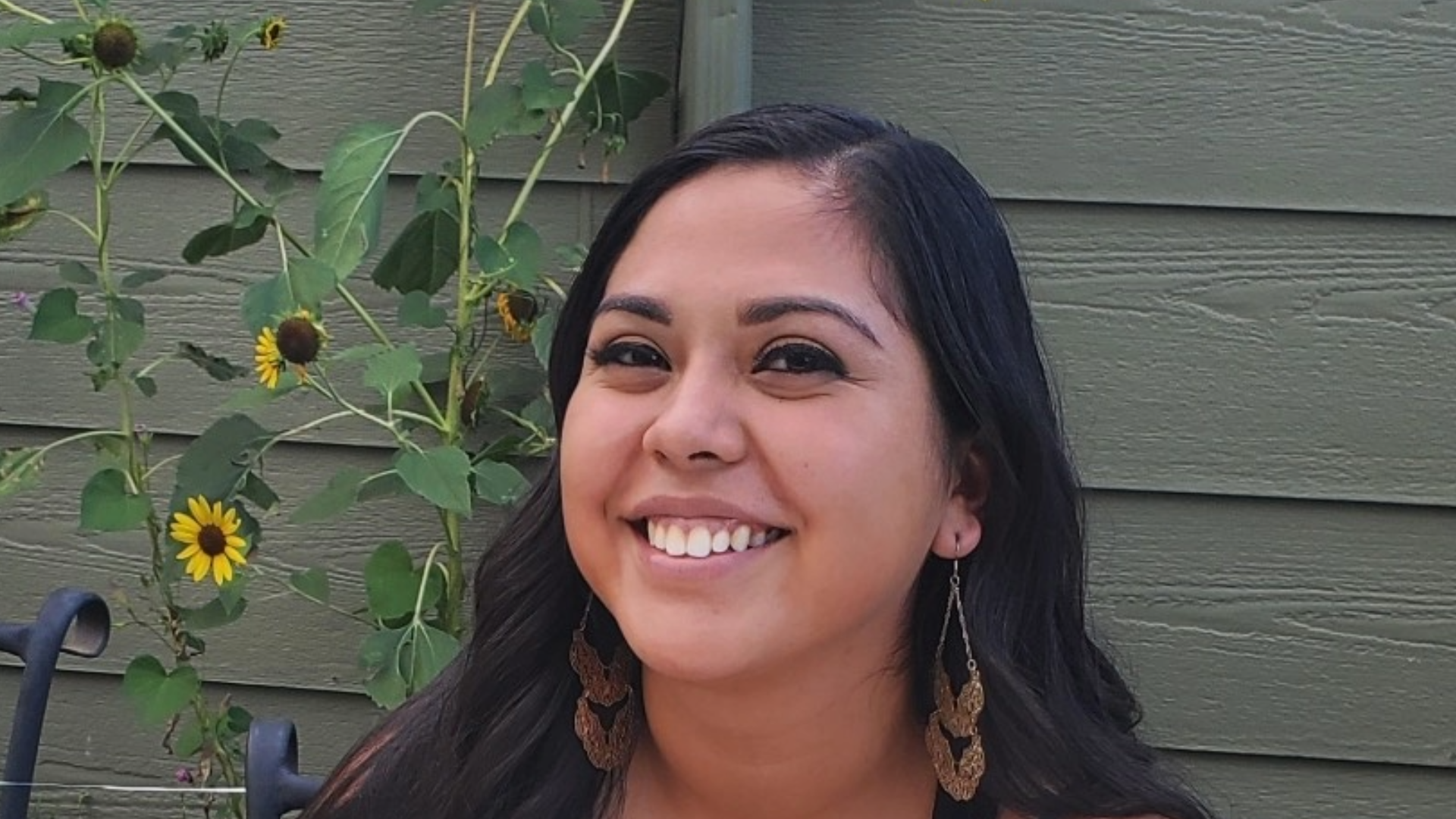 Sheena Leal with long, dark hair and a bright smile sits on a bench outdoors. She is wearing a black tank top with a lace detail and long, brown, patterned earrings. Behind her is a light green wall and several tall sunflower plants with yellow blooms.
