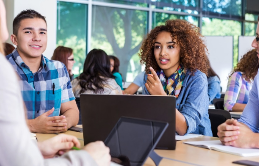 people sit at a table indoors speaking to each other with a laptop on the table